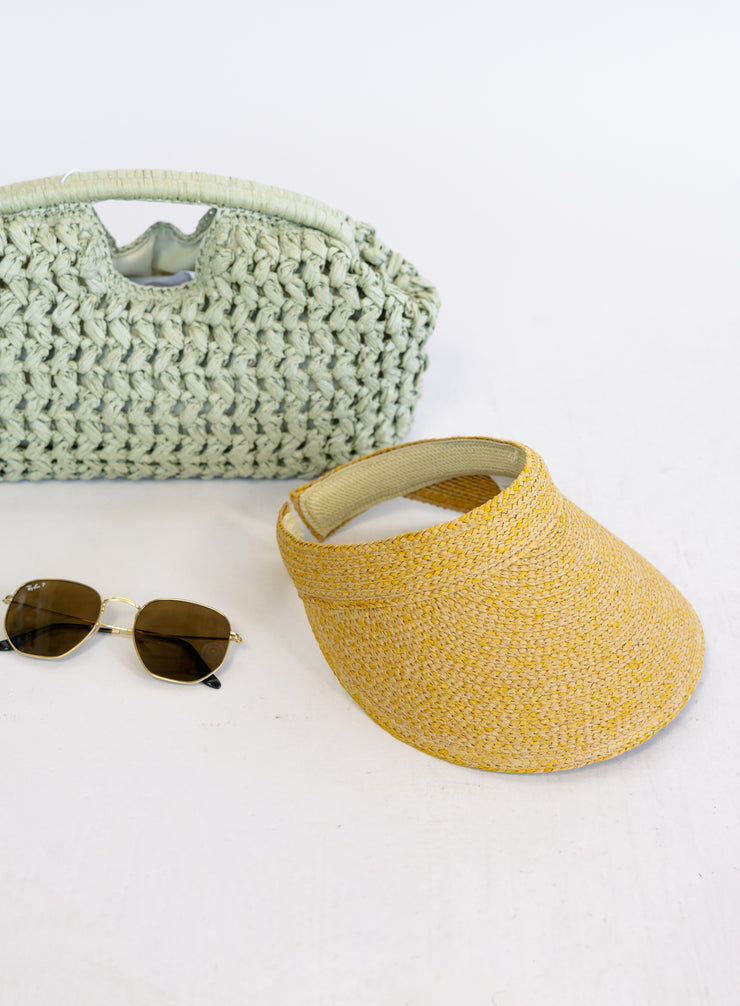 Flat lay of a woven bag, a straw visor and sunglasses